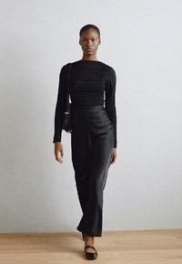 Schwarzes langärmeliges Kleid mit enganliegendem Oberteil und Raffung, gerader Silhouette und glatter, satinähnlicher Textur, kombiniert mit flachen schwarzen Schuhen.