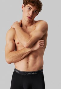 Crne Calvin Klein bokserice s elastičnom trakom s logom. Model ima kratku, kovrčavu kosu i bez majice je, pokazujući torzo i položaj ruku.