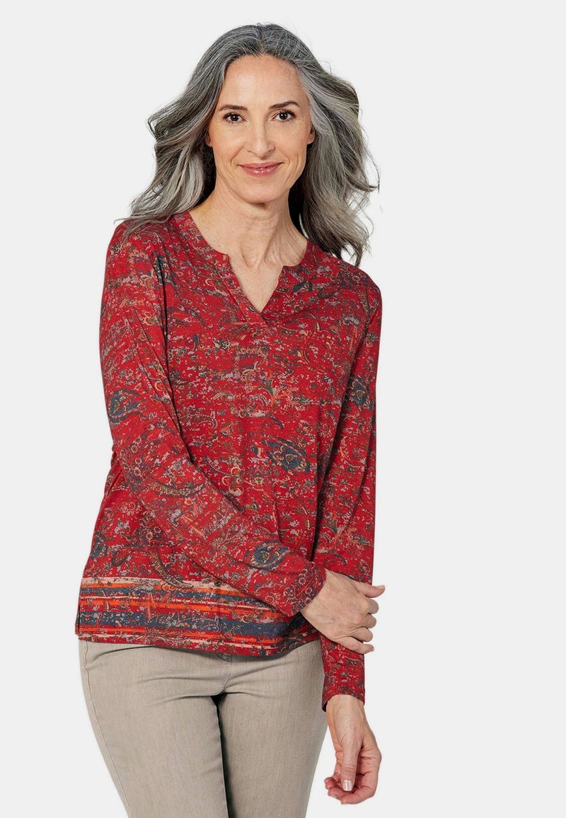 Rote Bluse mit langen Ärmeln, die ein Paisleymuster in Blau und Gold aufweist, mit einem ausgeschnittenen Ausschnitt und einem unifarbenen, gesäumten Saum.
