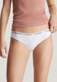 Slip bikini bianco in cotone con un cinturino grigio e nero riportante il marchio, texture liscia e taglio classico. Indossato con una maglietta aderente.