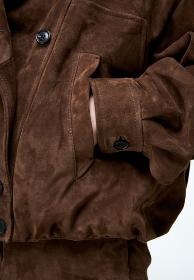 Hand met een bruine suède jas met een met knopen gesloten manchetkant, hand deels in de zijzak, close-up van stoftextuur en stiksel.
