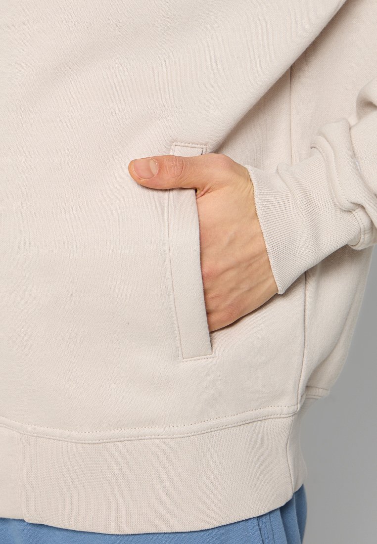 Mano inserita nella tasca anteriore di una felpa beige chiaro con polsini a costine, abbinata a pantaloni blu.