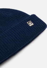 Gorro de malha azul-marinho com um padrão texturizado, com a dobra na parte de cima e uma pequena etiqueta retangular com detalhes em dourado.