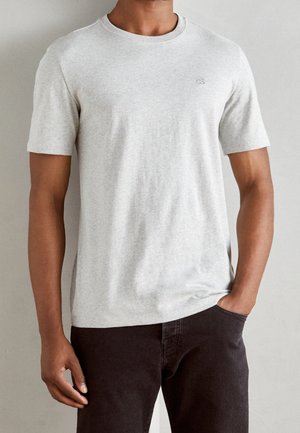 T-shirt gris clair à manches courtes, col rond, en tissu doux, avec un petit logo brodé sur la poitrine, associé à un pantalon foncé.