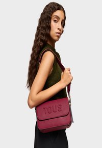 Bolso de bandolera de cuero burdeos con el logo "TOUS" grabado, diseño plano, correa ajustable y un acabado texturizado.