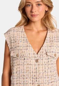 Beige strukturiertes Weste mit tiefem V-Ausschnitt, zwei Brusttaschen, goldenen Knöpfen und einem gewebten Muster mit blauen und braunen Akzenten.