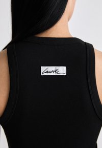 Frau trägt ein schwarzes, geripptes Tanktop mit einem weißen rechteckigen Etikett mit der Aufschrift "Lavote", das auf dem oberen Rücken angenäht ist.