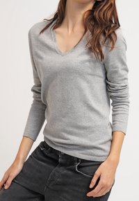 Pull-over gris en tricot avec un col en V, des manches longues et une silhouette ajustée. Le tissu semble doux et lisse, sans motifs visibles.