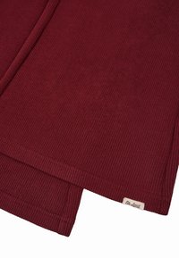 Bordeaux gebreid kledingstuk met ribbeltextuur, voorzien van een klein label in de rechterbenedenhoek. Rechthoekige vorm, zacht materiaal.