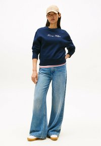 Felpa blu navy con ricamo "Tommy Hilfiger", abbinata a jeans chiari a gamba larga e un cappellino beige. Scarpe bianche con suola spessa.