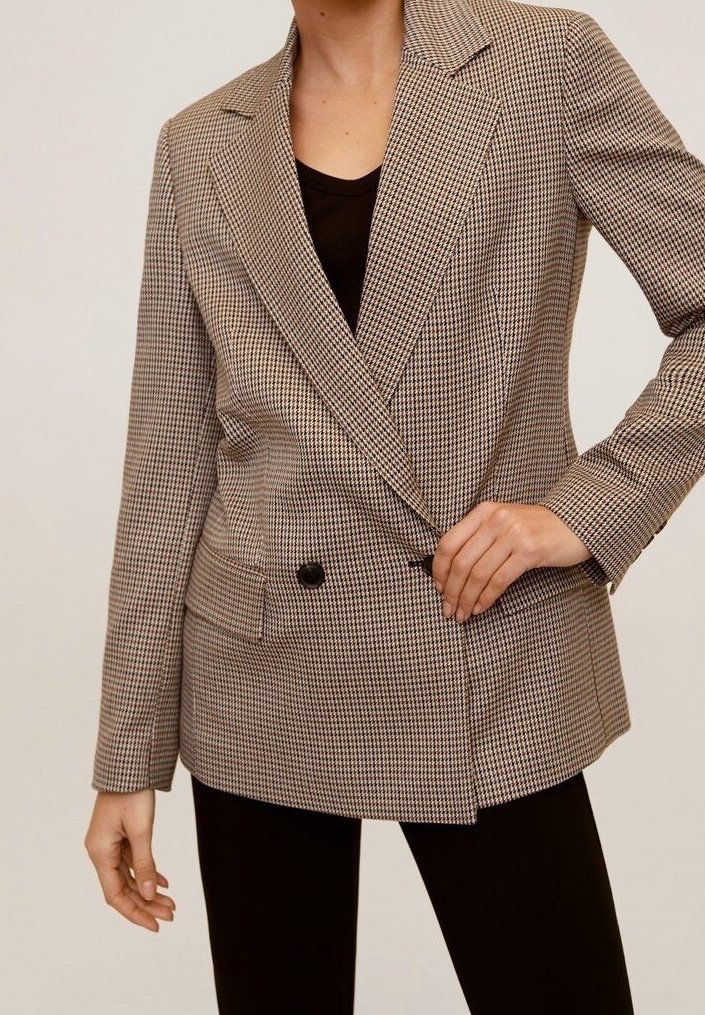 Femme portant un blazer à carreaux à double boutonnage sur un top noir et un pantalon noir, ajustant le bouton du blazer avec la main droite.
