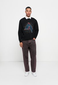 Black sweater with a blue and red graphic design, paired with dark purple jeans and white sneakers. Model standing against a plain background.