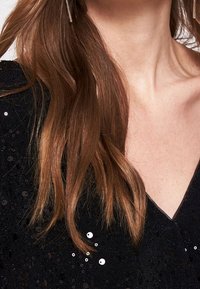 Top negro con lentejuelas y un escote en V profundo, que presenta tela texturizada y una superficie brillante, complementado con cabello largo y ondulado de color castaño.