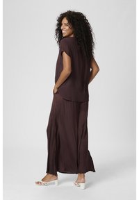 Une tenue longue et fluide de couleur bordeaux profond, avec des manches courtes et des fentes sur les côtés. Portée avec des sandales à talons carrés blanches et à lanières.