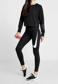 Svart kort sweatshirt med en logotyp, kombinerad med svarta leggings med en vit swoosh. Svarta sportskor fulländar outfiten. Håller en rosa flaska.
