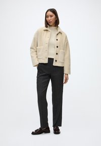 Šviesiai ruda pūkinė striukė su apykakle, priekiniais mygtukais ir dviem kišenėmis, derinama su tamsiomis tekstūruotomis kelnėmis ir tamsiais batais.