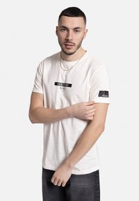T-shirt à manches courtes en crème avec une texture douce et un logo noir imprimé sur la poitrine et la manche ; associé à un jean foncé.