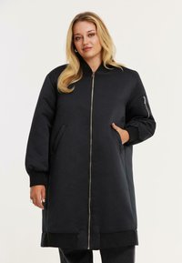 Vrouw met blond haar in een lange zwarte pufferjack met een ritssluiting aan de voorkant en handen in de zakken tegen een effen lichtgekleurde achtergrond.