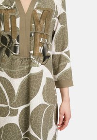 Lång tunikaklänning i olivgrön med djärv grafisk tryck av stora blad, med dekorativa metallbokstäver och V-ringning.