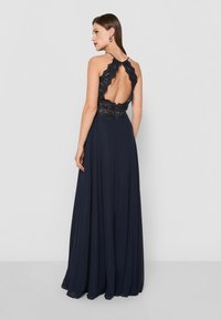 Tengerészkék estélyi ruha csipkebetéttel a háton, nyakpántos fazonnal, lebegő sifon szoknyával és bonyolult csipkés díszítéssel a vállaknál.