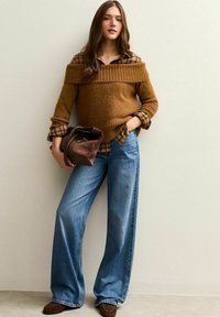 Bruine gebreide trui over een geruit overhemd, gecombineerd met blauw wijde pijpen spijkerbroek. Accessoires zijn een bruine handtas en leren schoenen.