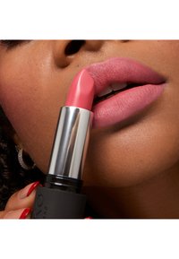 Un lápiz labial rosa con una textura suave sostenido cerca de los labios, encerrado en un tubo negro con una tapa plateada metálica. Acabado mate en los labios. Uñas rojas visibles.