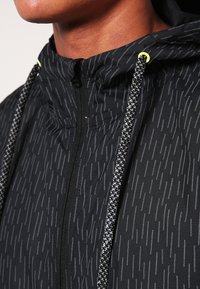 Svart huva jacka med en texturerad regnmönster. Har grå reflexdetaljer och dragkedja framtill. Dragsko med gula spetsar.