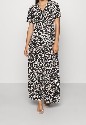 Mujer que lleva un vestido cruzado negro con patrones florales blancos y beige, mangas cortas y sandalias blancas abiertas de punta, de pie sobre un suelo gris.