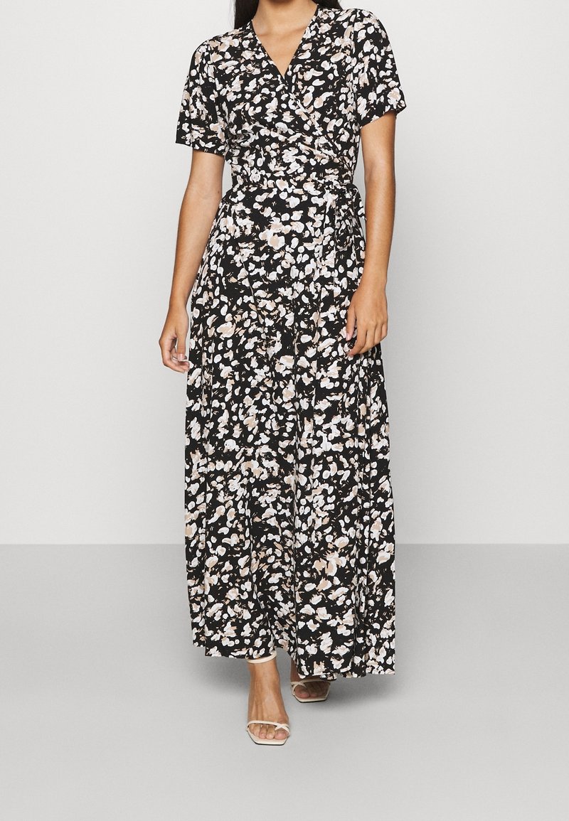 Femme portant une robe portefeuille noire avec des motifs floraux blancs et beiges, manches courtes, et des sandales blanches à bouts ouverts, debout sur un sol gris.