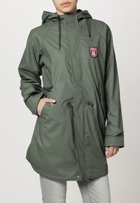 Personne portant un long imperméable vert olive avec une capuche, des cordons de serrage et des poches avant, debout devant un fond uni.