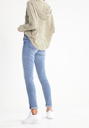 Person steht, trägt hellblaue Jeans, beige Kapuzenpullover und weiße Turnschuhe, und blickt mit dem Rücken zu einem weißen Hintergrund.