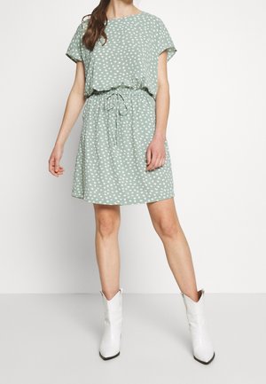 Donna che indossa un vestito verde chiaro a maniche corte con pois bianchi e vita annodata, abbinato a stivali bianchi fino a metà polpaccio, in piedi su un pavimento bianco.