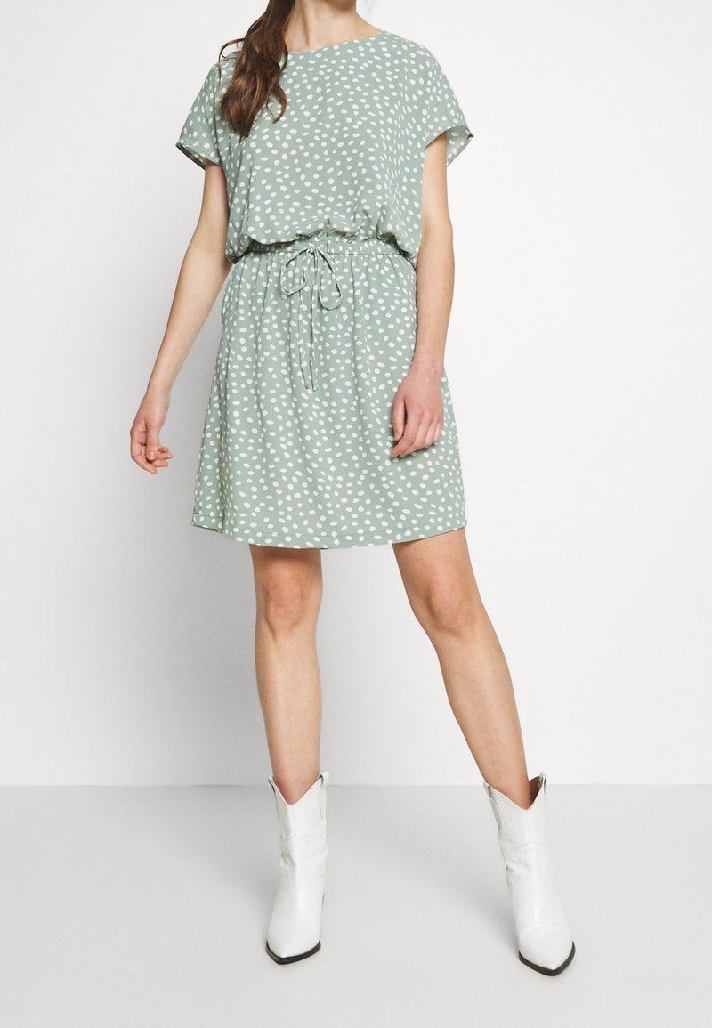 Femme portant une robe courte à manches courtes vert clair à pois blancs avec une taille nouée, associée à des bottes blanches mi-mollet, debout sur un sol blanc.