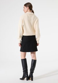 Suéter de canalé crema, falda corta negra y botas altas de cuero negro con tacones cuadrados. Diseño minimalista, textura suave.