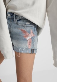 Shorts en denim de couleur bleu délavé, avec un motif d'oiseau rose brodé sur la jambe gauche, un ourlet effiloché et des détails usés.