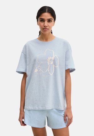 Kobieta w jasnoniebieskiej, dużej koszulce z dużym kwiatowym nadrukiem i pasujących szortach, stojąca na jednolitym tle.