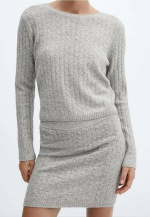 Vrouw draagt een lichtgrijze kabelgebreide trui met lange mouwen en een bijpassend gebreid minirokje, staand tegen een effen achtergrond.