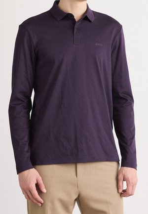 Mann trägt ein dunkelviolettes langärmeliges Polohemd mit beiger Hose, steht mit entspannten Händen seitlich vor einem hellen Hintergrund.