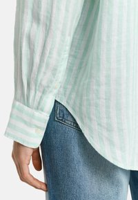 Camisa de manga larga ligera a rayas en verde menta y blanco con puños abotonados; dobladillo ligeramente curvado; combinada con pantalones vaqueros de mezclilla azul.
