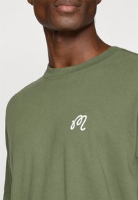 Olīvu zaļas kokvilnas T-krekls ar apaļu kakla izgriezumu un īsām piedurknēm. Uz krūtīm ir balts kursīvs logo. Mīksta tekstūra.