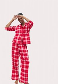 Czerwony zestaw piżamowy w kratę, składający się z koszuli zapinanej na guziki z kieszenią na piersi oraz luźnych spodni. Materiał ma gładką fakturę z kontrastującymi różowymi liniami.