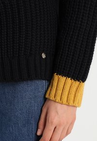 Gruby czarny sweter z dzianiny z prążkowanym musztardowym ściągaczem i małym metalicznym logo z boku. Miękka tekstura i luźny krój.