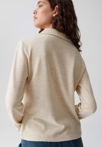Woman with long curly hair wearing a beige knit sweater with a ribbed collar, standing with hands in pockets, facing away from the camera.