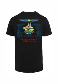 Svart t-skjorte med grafisk design av en hundelignende figur med gule og blå striper, omkranset av navn på land, og teksten "VERDENSLAVETUR 1984-85".