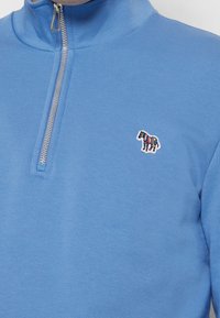 Maglione azzurro chiaro con cerniera a quarto di lunghezza argento e piccolo logo zebra multicolore ricamato sul petto a sinistra.