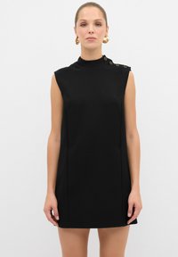 Vestido negro sin mangas con un escote alto, que presenta un detalle de botones en el lateral y una silueta estilizada. Tejido suave, hasta la rodilla.