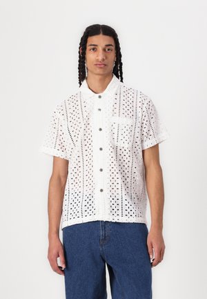 White short-sleeve shirt made of embroidered fabric with cut-out patterns, featuring a pointed collar, button closure, and a chest pocket.