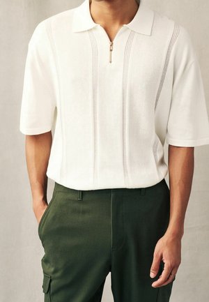 Homme portant un polo en maille blanc à manches courtes avec fermeture éclair et pantalon vert foncé, mains dans les poches, fond neutre.