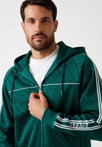 Sudadera verde con cremallera y acentos blancos, que presenta un patrón repetido, capucha con cordón y detalles de marca en las mangas. Tejido texturizado.
