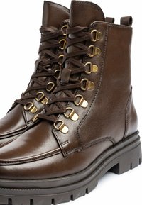 Bottines en cuir marron avec des garnitures dorées, présentant un design robuste à lacets, une finition texturée et une semelle épaisse et rugged.